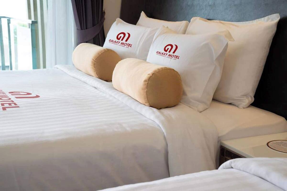 Una cama bien hecha con ropa de cama blanca y dos almohadas color beige, una almohada blanca con el logo del Galaxy Motel y otra almohada blanca con el logo del Galaxy Motel.