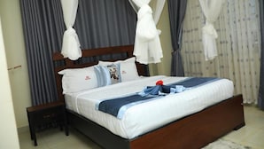 Una cama bien hecha con sábanas blancas, una manta a rayas azul y blanco, y una decoración floral roja. Junto a la cama hay una mesita de noche de madera, y la habitación tiene cortinas grises y un dosel blanco sobre la cama.
