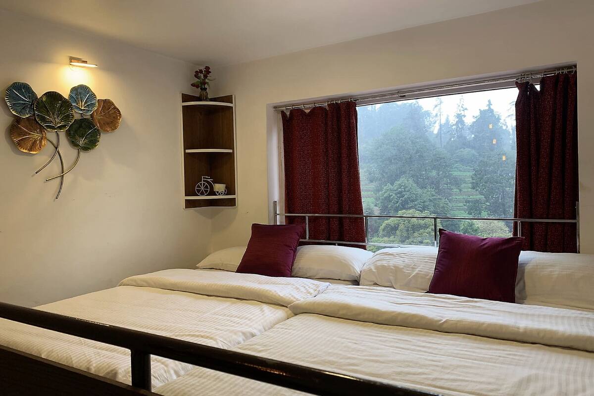 A bedroom with a bed, a window with curtains, a wall-mounted shelf with decorative items, and a view of a forest through the window.