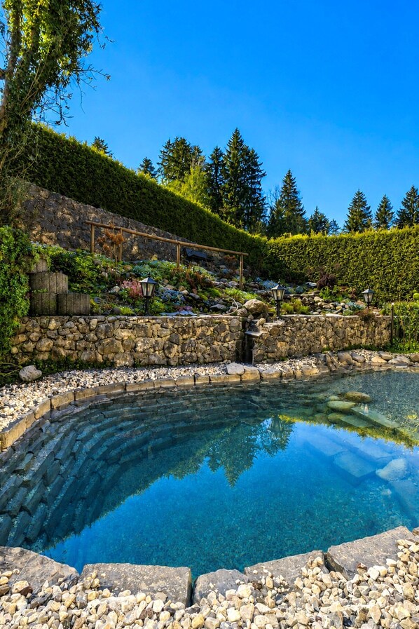Ein klares Schwimmbecken, umgeben von einer Steinmauer, mit einer Holzterrasse und einer Steintreppe, die zu einer Holzpergola führt.