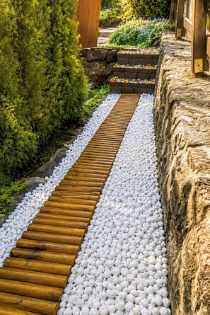 Ein Weg mit Holzplanken und weißen Kieselsteinen, umgeben von Grünpflanzen und einer Steinmauer.