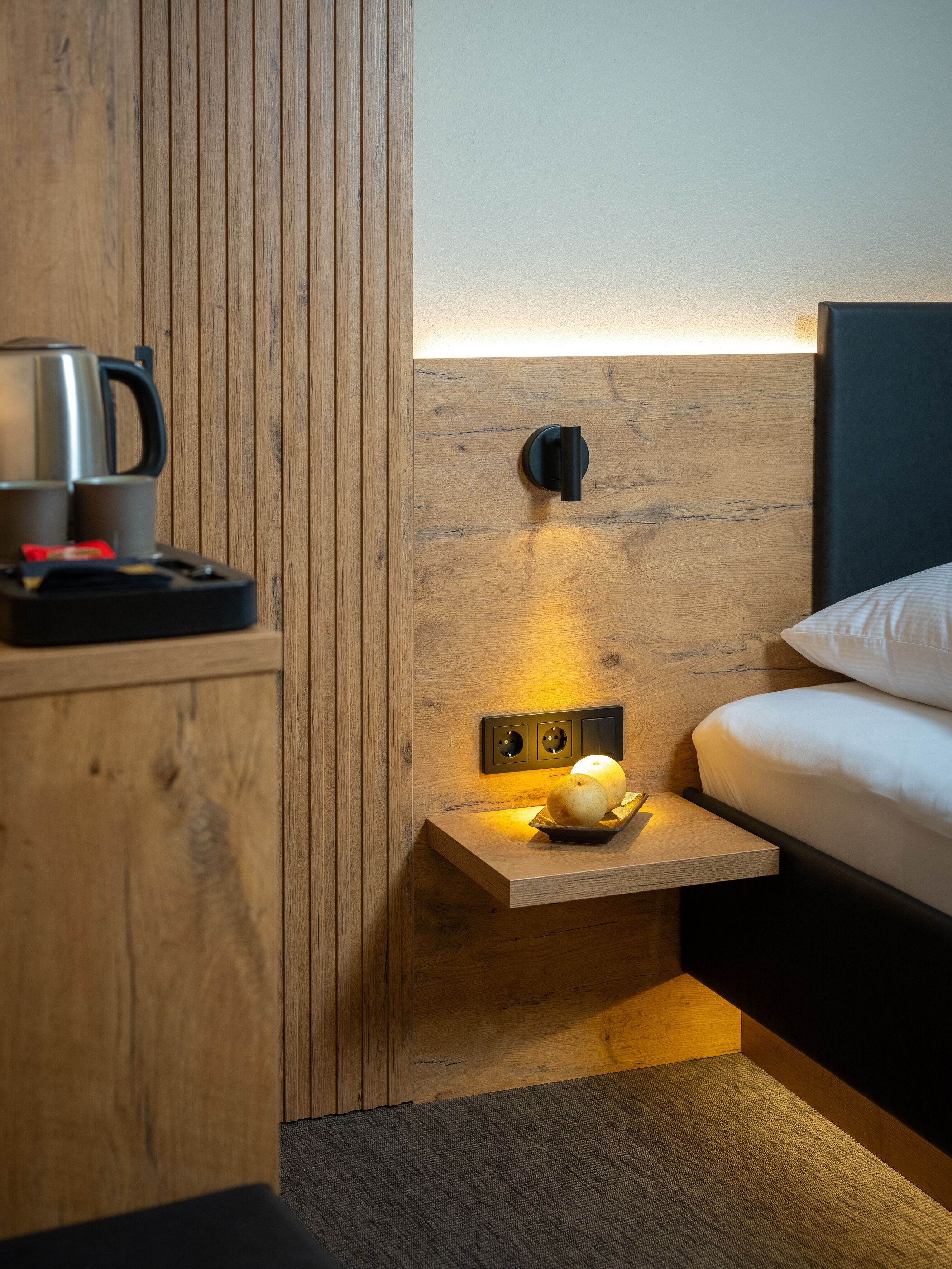 Een kleine hotelkamer met een wand van houten panelen, een nachtkastje met een lamp, een waterkoker en een bord met twee appels.