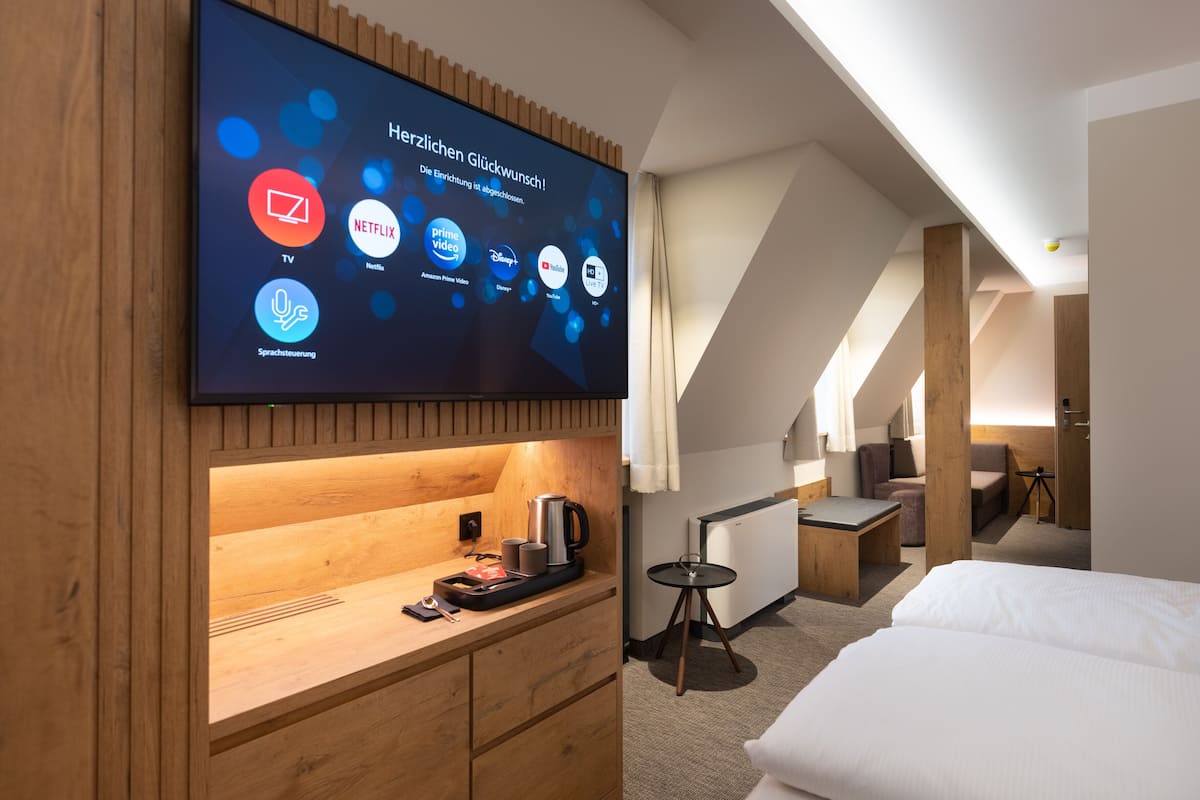 A modern hotel room with a flat-screen TV displaying streaming services, a wooden desk with a coffee maker, a bed, and a small table with a lamp.