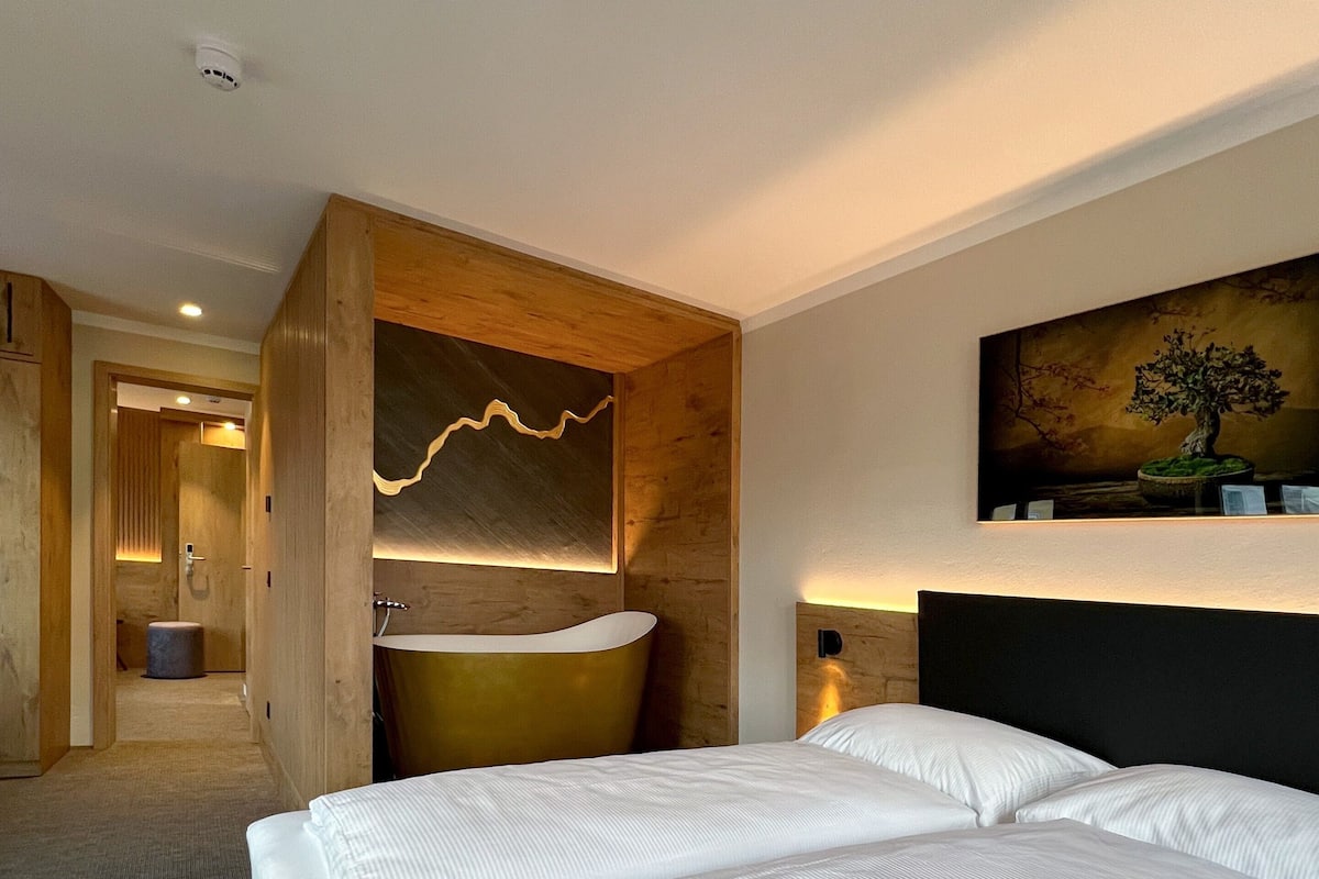 A hotel room with a large bed, a wooden accent wall, a round bathtub, and a painting of a tree.
