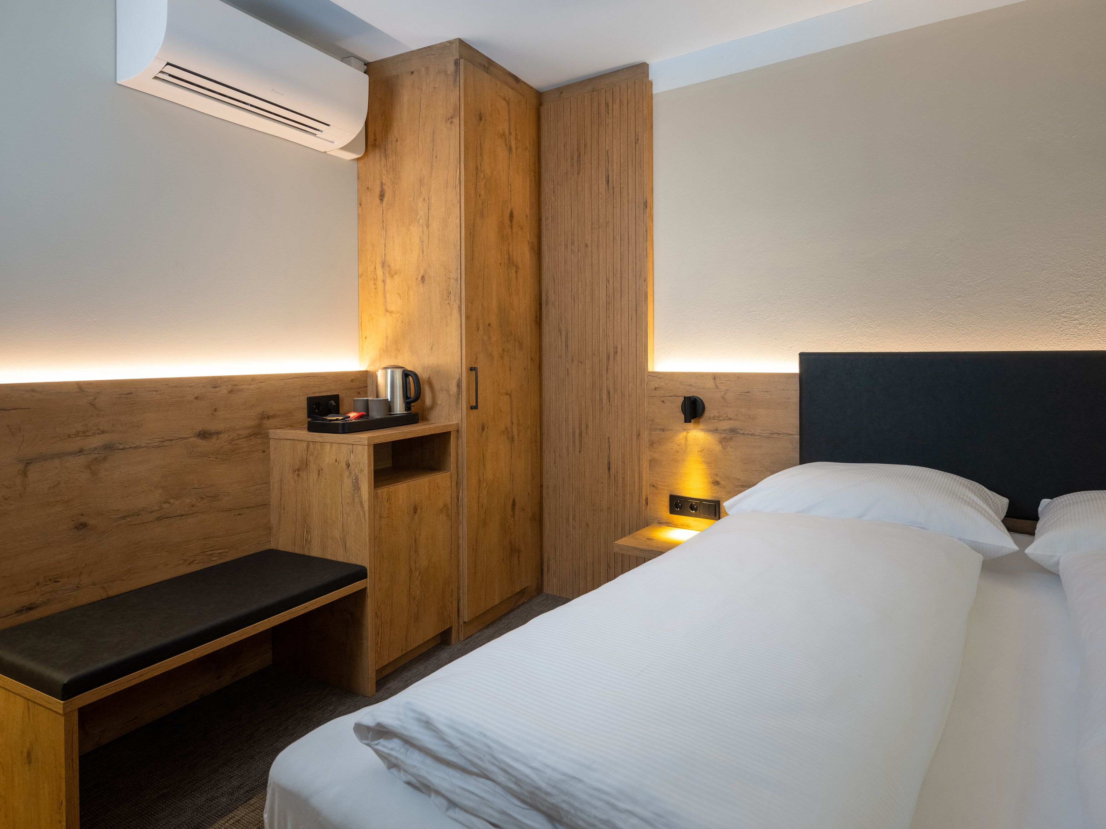 A hotel room with a bed, a bench, a wardrobe, and a bedside table with a kettle and cups.