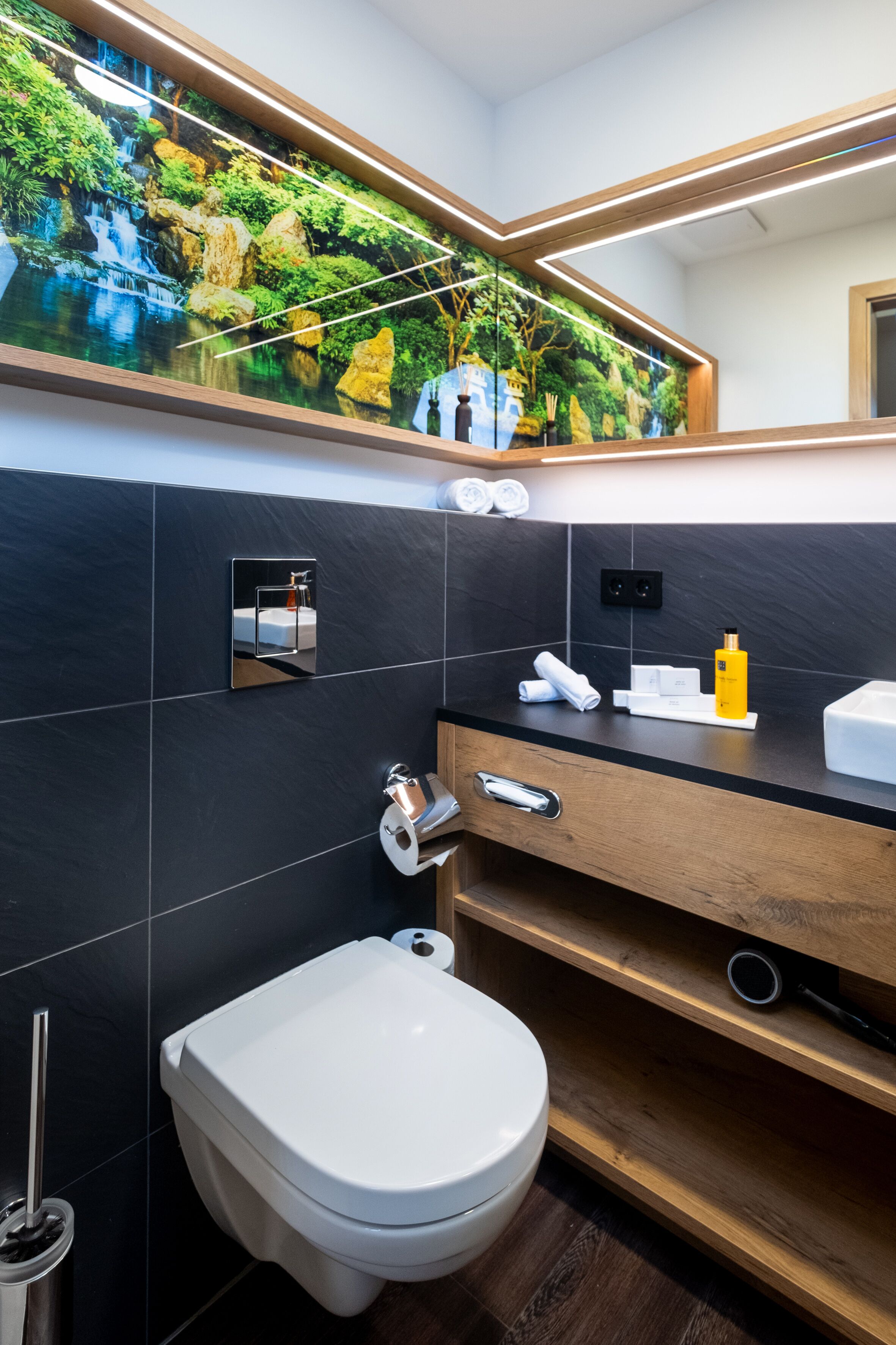 Casa de banho moderna com parede revestida a azulejos pretos, sanita branca, toucador de madeira e espelho com painel decorado com motivos naturais.