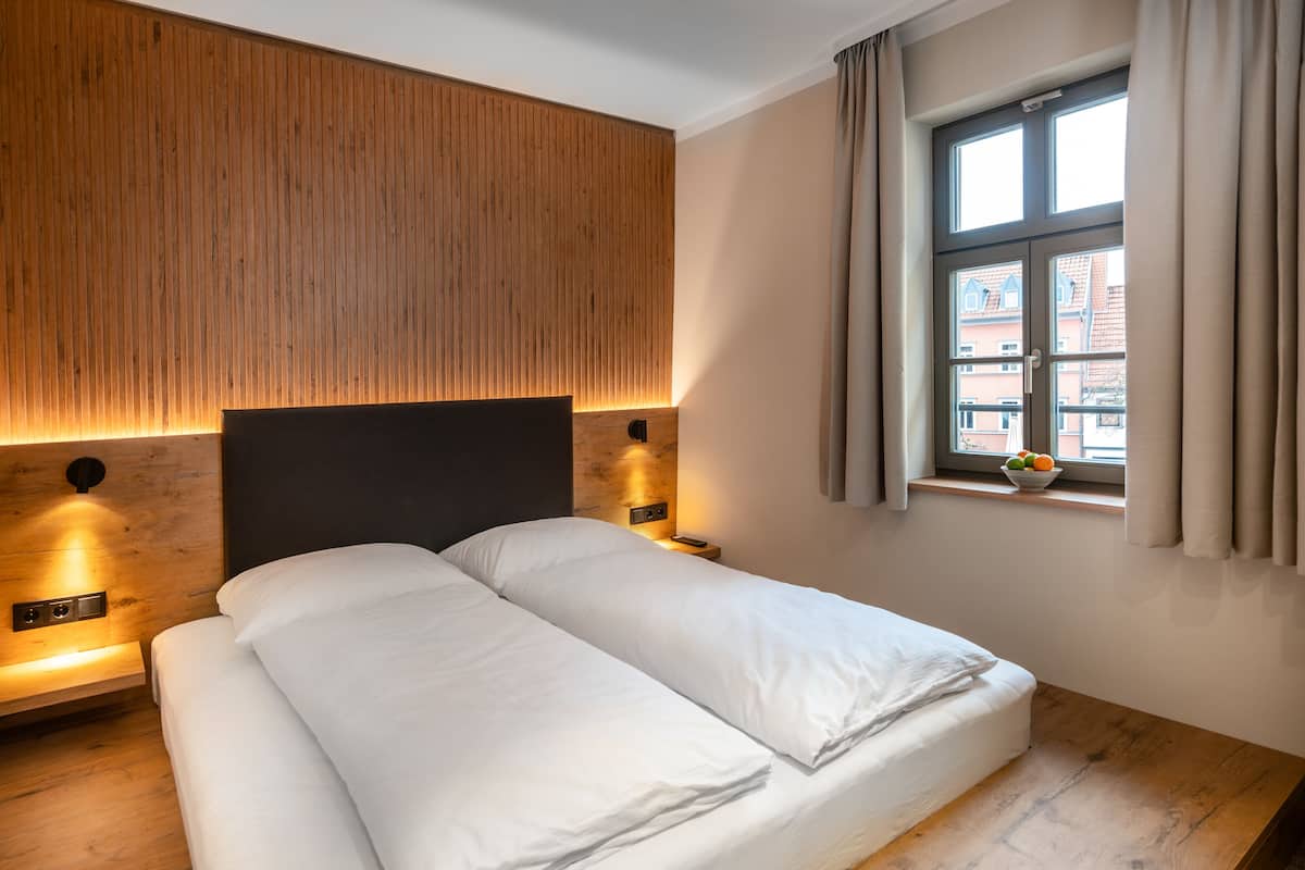 A modern bedroom with a wooden headboard, a large bed, bedside lamps, and a window with curtains.