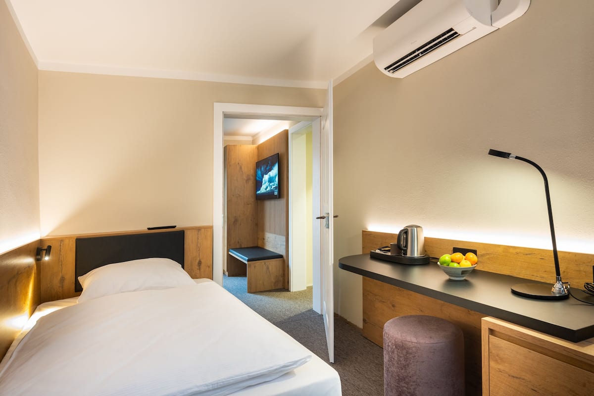 A hotel room with a bed, a desk with a kettle and fruit, a TV, and an air conditioning unit.