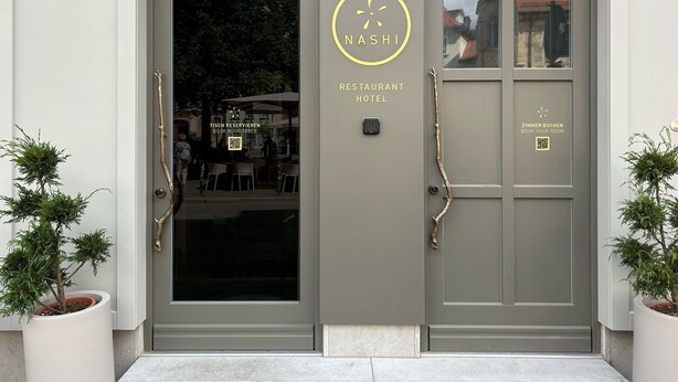 En hotellentrén med modern design, med en glasdörr och en skylt som visar texten "NASH RESTAURANT HOTEL". Här finns QR-koder för bords- och rumsbokning, och ett litet fönster som visar interiören.