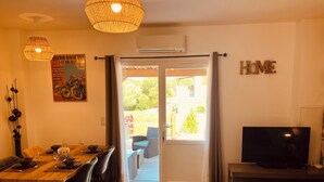 Un comedor con mesa y sillas de madera, un televisor sobre un mueble bajo, y un toldo de vidrio corredizo que da a un patio exterior.