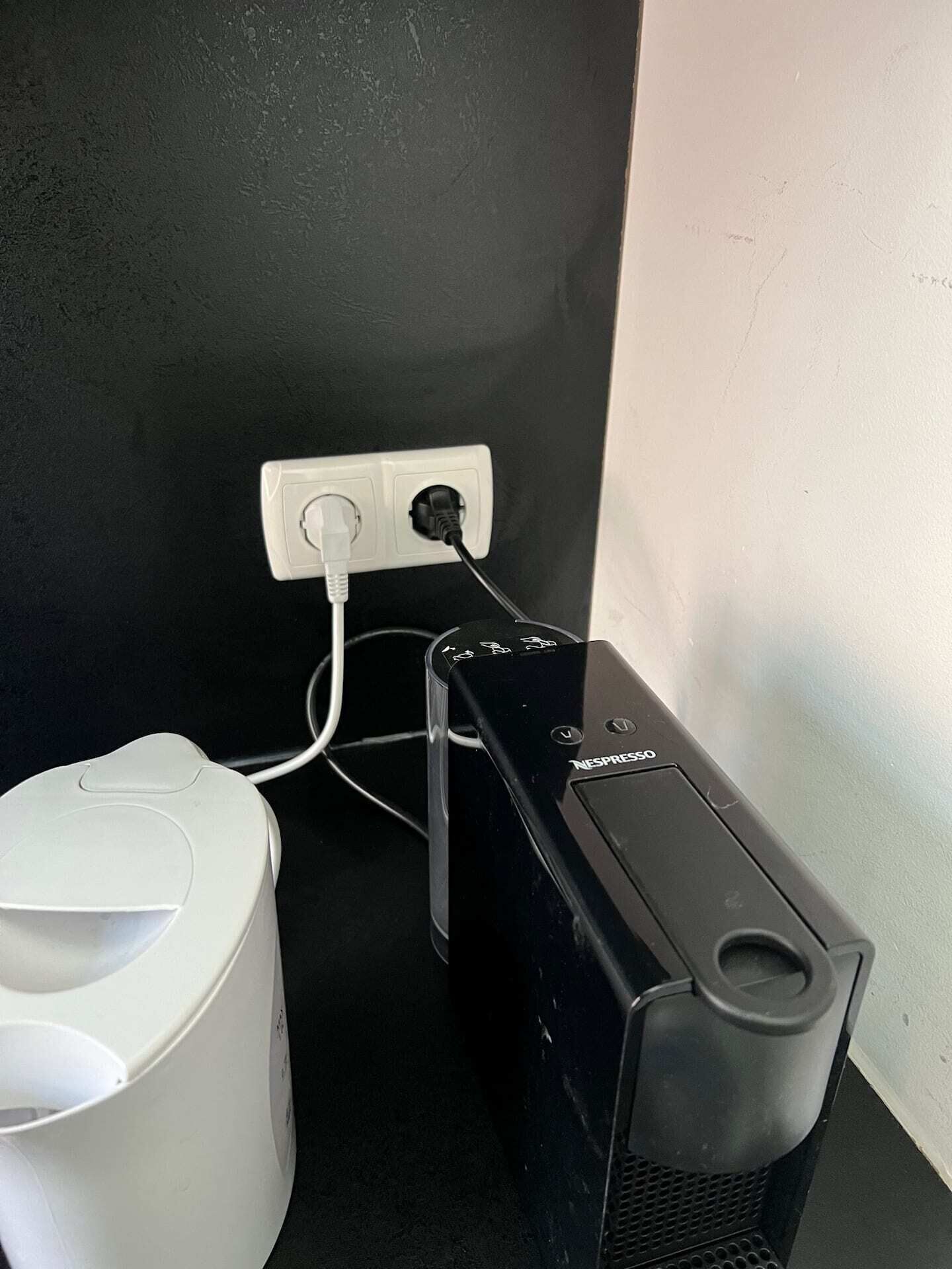 A black Nespresso coffee machine connected to a white power outlet, with a white coffee maker on a black countertop.