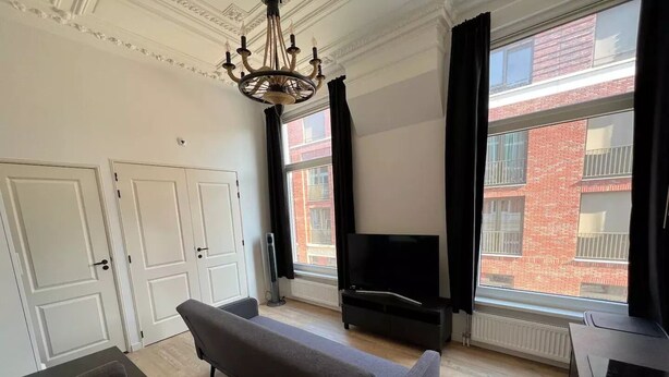 Une pièce avec un banc, un téléviseur, un radiateur et une fenêtre donnant sur un immeuble en briques rouges.