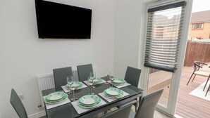 Et spiseområde med et bord dækket til seks personer, et fladskærms-tv på væggen og en skydedør til en udendørs terrasse.