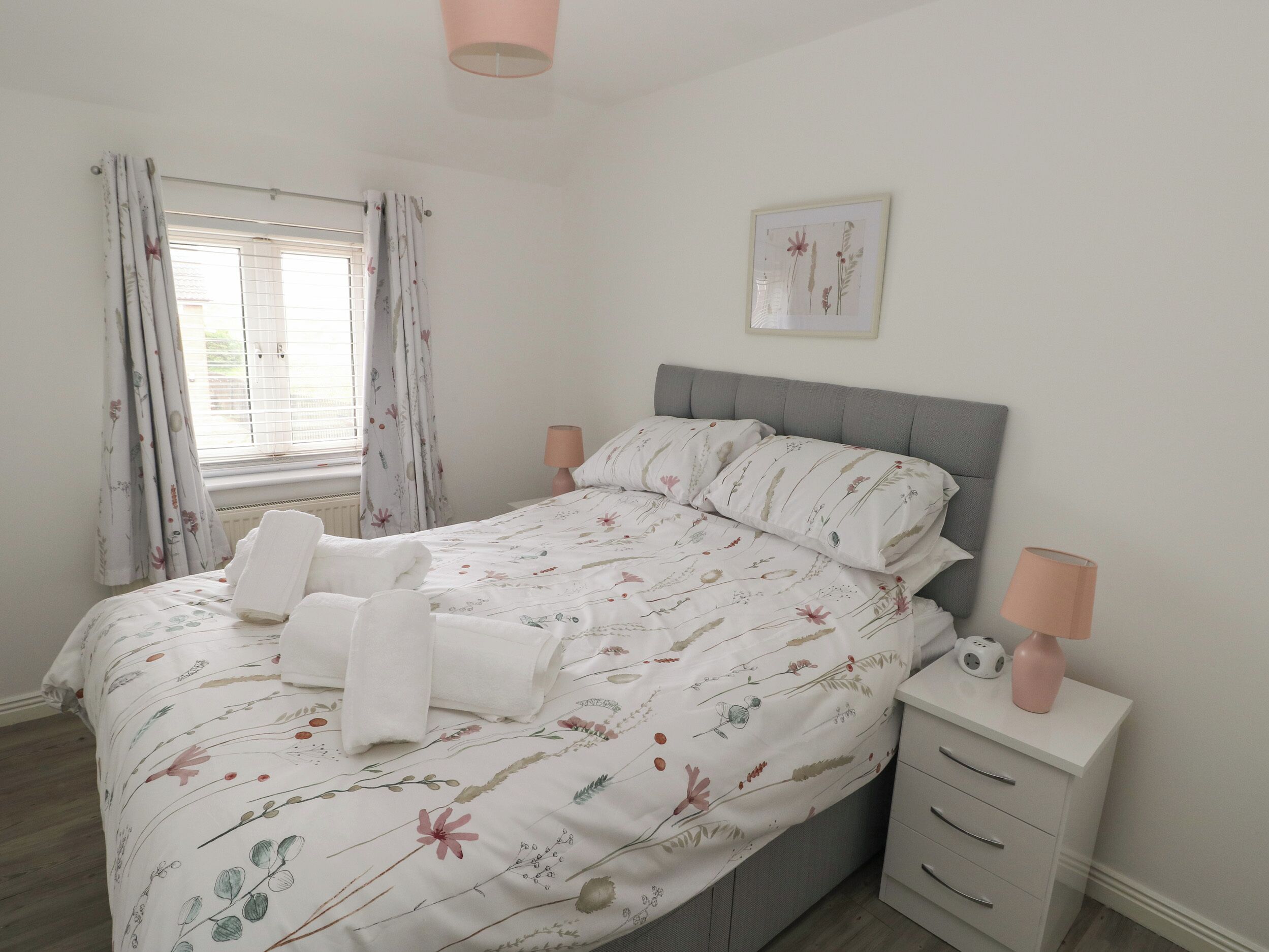 Una cama bien hecha con ropa de cama floral, una lámpara de noche y una mesita de noche pequeña con un florero.