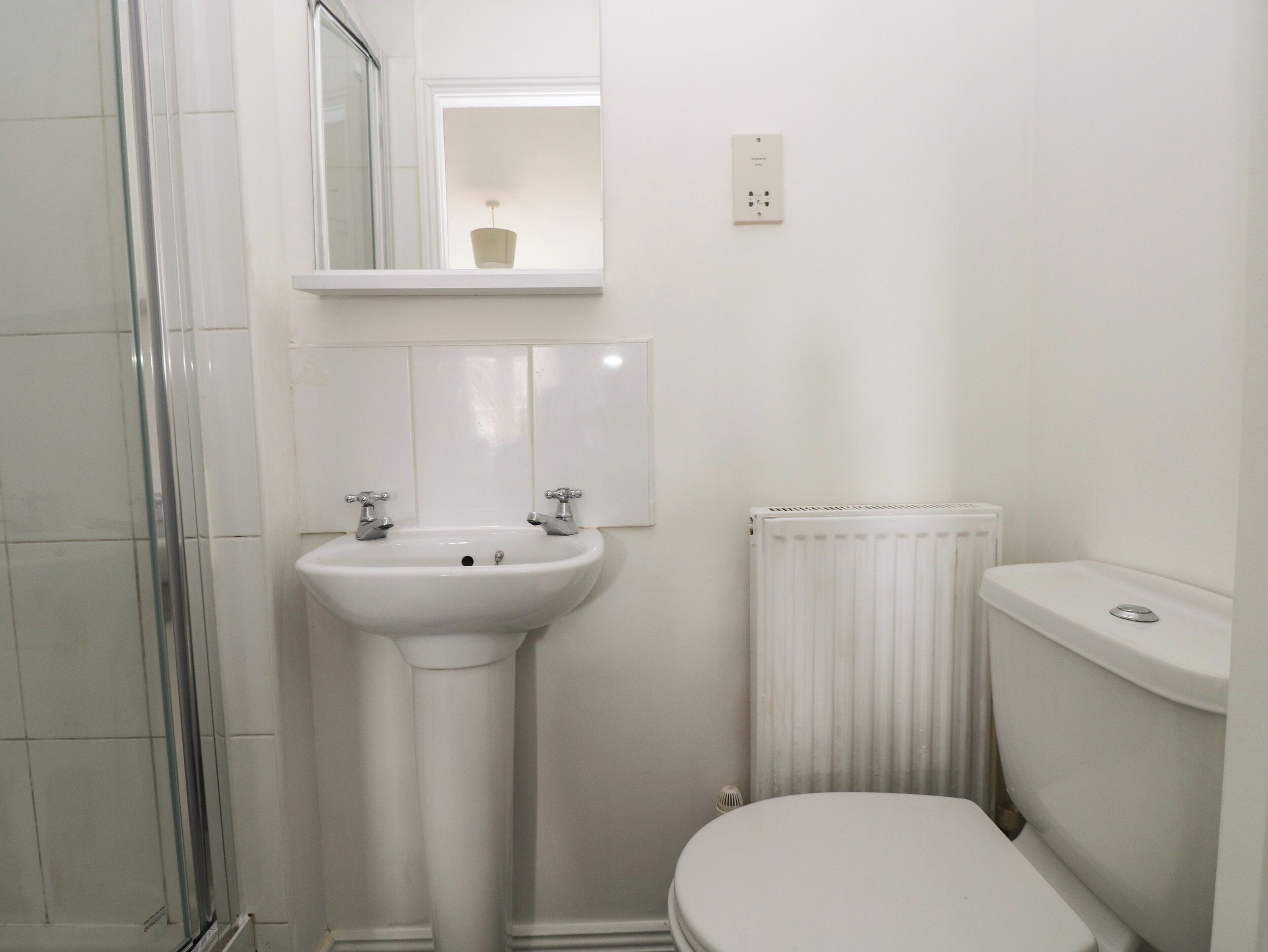 Un baño con lavamanos pedestal, inodoro, radiador y cabina de ducha con mampara de vidrio.