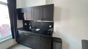 Cocina compacta con armarios oscuros, encimera negra, un lavamanos y un cubo de basura.
