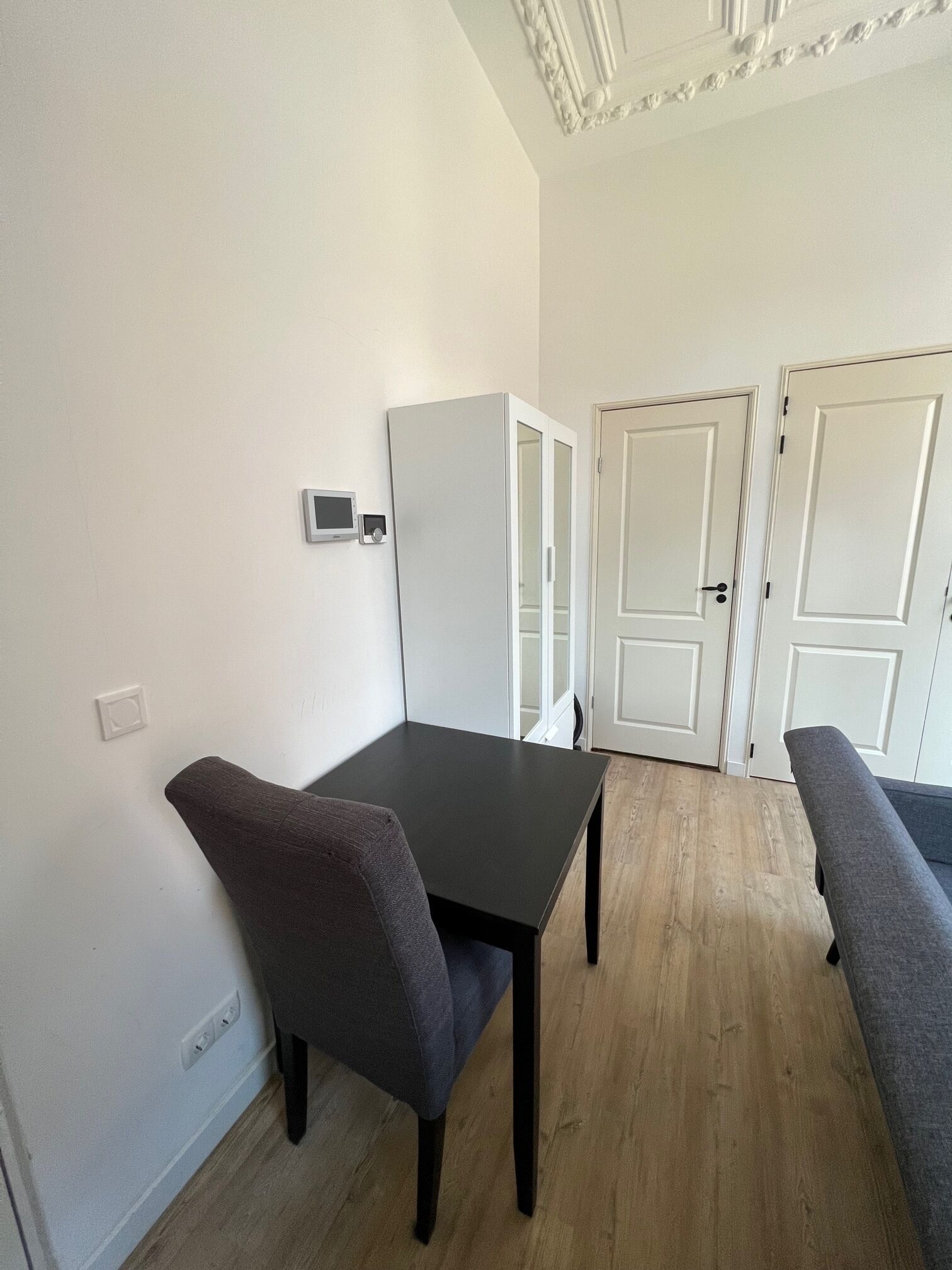 Una habitación con una mesa de madera oscura, dos sillas grises, un armario blanco y una puerta blanca.