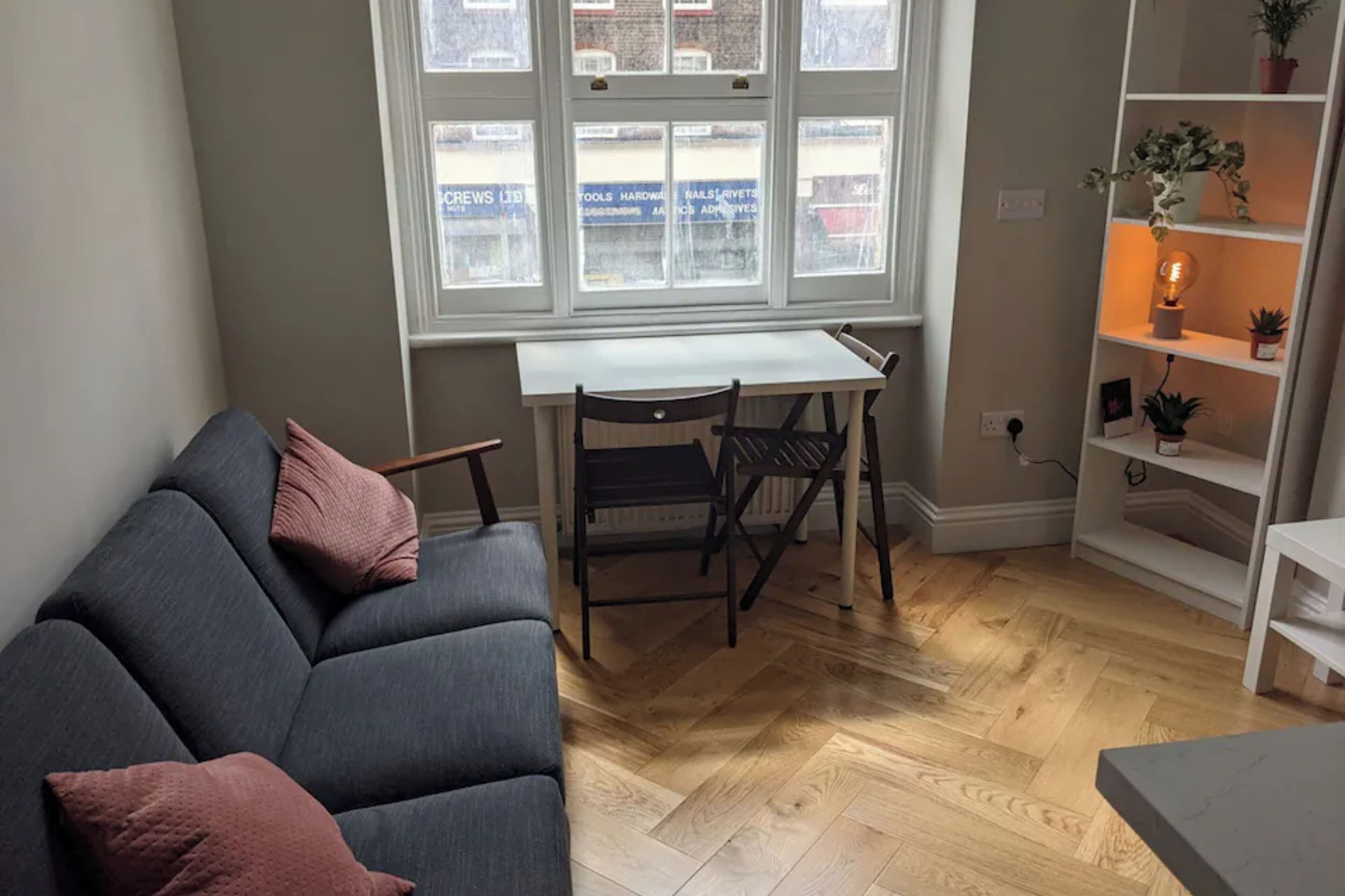 A room with a grey sofa, a small table, and chairs. There is a window with a view of buildings outside.