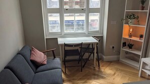 Une pièce avec un canapé gris, une petite table et des chaises. Il y a une fenêtre donnant sur des bâtiments à l’extérieur.