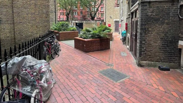 Une ruelle étroite pavée de briques, où l’on aperçoit un vélo recouvert de plastique, une clôture en métal et un jardin en briques orné de plantes vertes.
