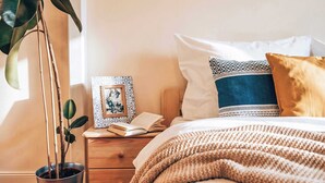 Ein Schlafzimmer mit einem Bett, Kissen, einem Nachttisch, einem gerahmten Bild, einer Topfpflanze und einer gestrickten Decke.