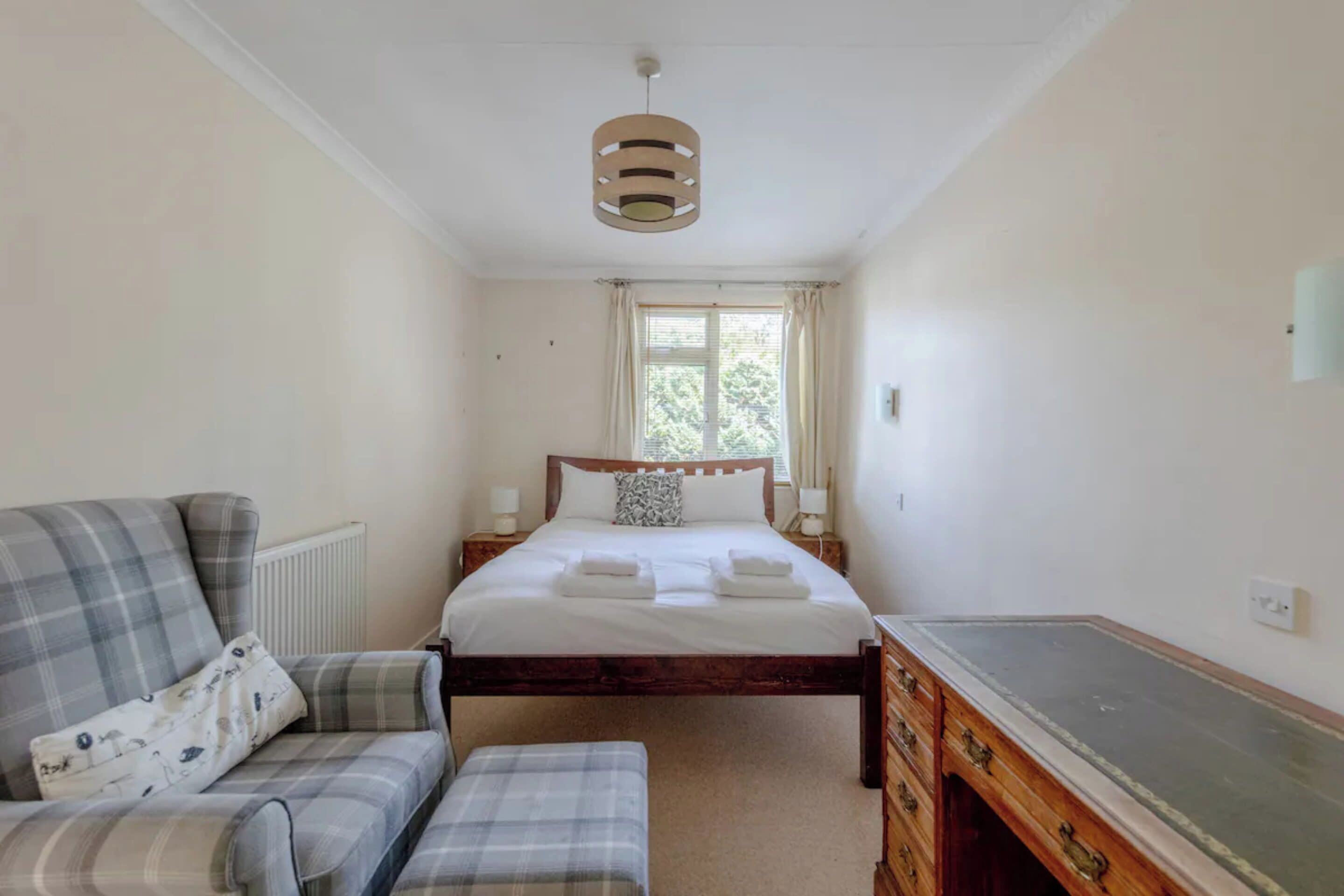 A bedroom with a bed, a wooden dresser, a bench, a radiator, a window with curtains, and a hanging light fixture.