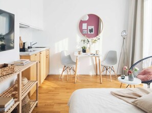 A modern, well-lit room with a bed, a desk with two chairs, a round mirror, and a small table with a lamp.