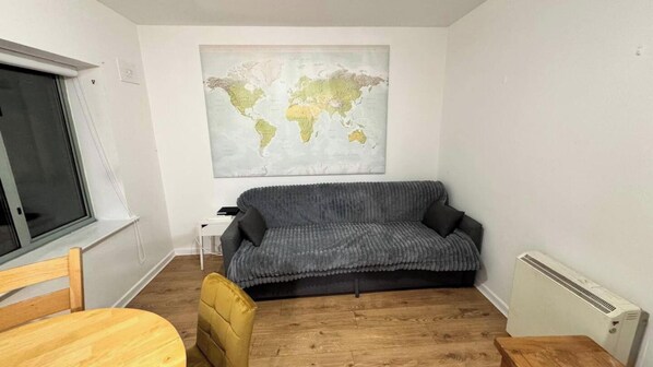 Een kleine, gemeubileerde kamer met een grijze bank, een houten stoel, een klein tafeltje, een wereldkaart aan de muur en een raam.