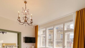A modern living room with a sofa, two armchairs, a coffee table, and a TV. There is a large window with curtains, a chandelier, and a patterned rug. - TurnKey | Vintage 2BR Apartment with Balcony (Praha)