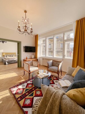 A modern living room with a sofa, two armchairs, a coffee table, and a TV. There is a large window with curtains, a chandelier, and a patterned rug. - TurnKey | Vintage 2BR Apartment with Balcony (Praha)