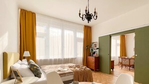 A hotel room with a bed, a sitting area with a table and chairs, a bookshelf, and a view through a window. - TurnKey | Vintage 2BR Apartment with Balcony (Praha)