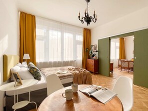 A hotel room with a bed, a sitting area with a table and chairs, a bookshelf, and a view through a window. - TurnKey | Vintage 2BR Apartment with Balcony (Praha)