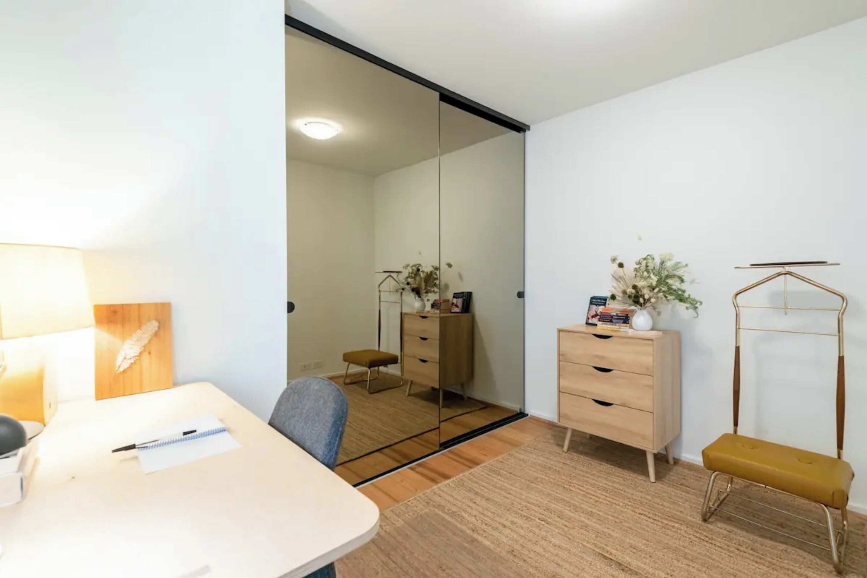 Una habitación moderna con escritorio, silla y espejo. Incluye un aparador de madera y una banca.
