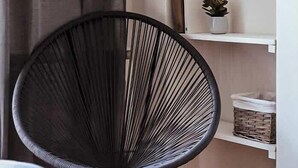 A room with a black wire chair, a white bookshelf with books and plants, and a grey curtain.