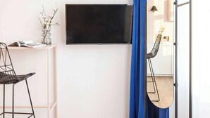 Habitación con televisor de pantalla plana en la pared, una pequeña mesa blanca con una revista y un jarrón de flores secas, una silla de metal negro y un espejo grande con marco de madera.