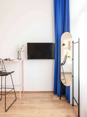 A room with a flat-screen TV mounted on the wall, a small white table with a magazine and a vase of dried flowers, a black metal chair, and a large mirror with a wooden frame.