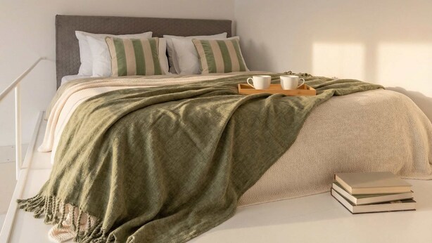 A neatly made bed with pillows, a green throw blanket, and a tray with two cups.