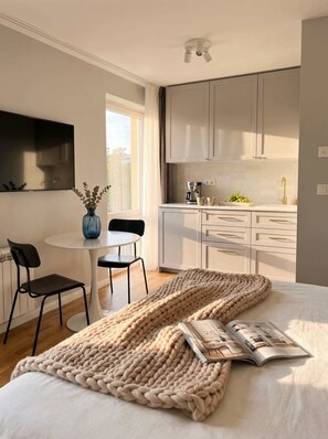 Un dormitorio moderno con una cama grande, una mesita de noche con una revista, una ventana con cortinas y una zona de cocina con armarios y una cafetera.