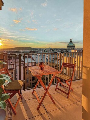 Un balcón con mesa y sillas, con vistas a un paisaje urbano con edificios y una estructura en forma de cúpula.