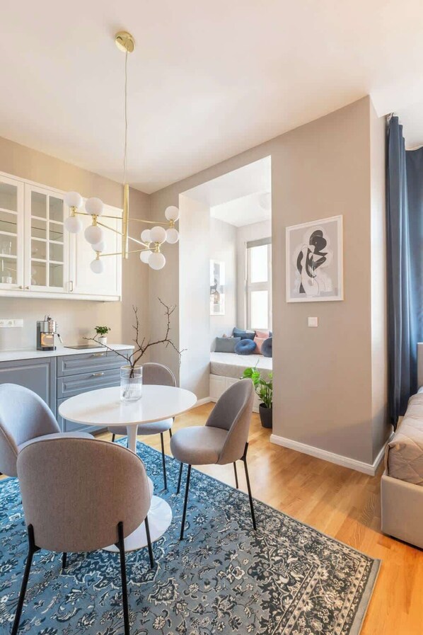 A modern living room with a round dining table, grey chairs, a blue patterned rug, a hanging light fixture, and a framed artwork on the wall.