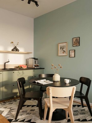 A modern kitchen with a round dining table, black chairs, a white vase with dried flowers, and a coffee machine.