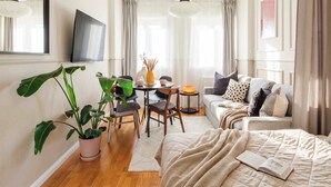 A modern hotel room with a flat-screen TV, a dining area with a table and chairs, a sofa with cushions, and a large potted plant.