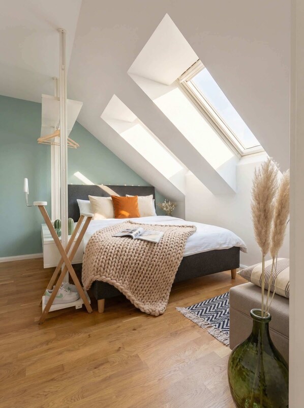 A bedroom with a skylight, a bed with pillows, a bedside table, a hanging light, and a vase with pampas grass.