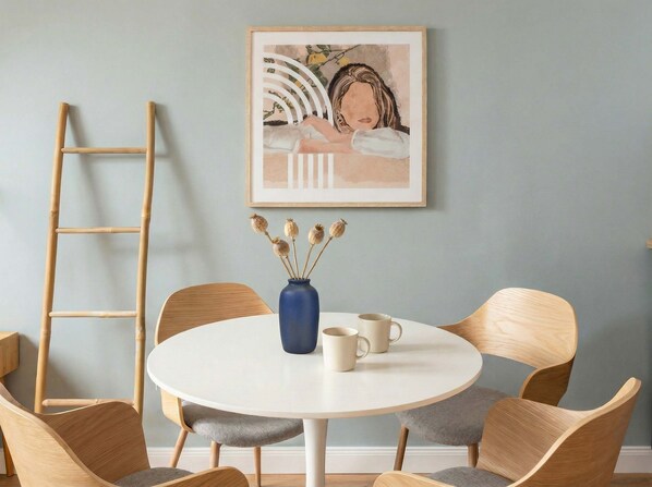 Ein runder Tisch mit Stühlen, eine Vase mit getrockneten Blumen und ein gerahmtes Kunstwerk an der Wand.