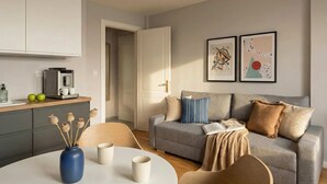 A modern living room with a grey sofa, a coffee machine, a vase with dried flowers, and two framed artworks on the wall.