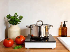 Eine Küche mit einem Edelstahlkochtopf auf einem Induktionskochfeld, einer Vase mit Grünpflanzen und zwei Tomaten auf der Arbeitsfläche.