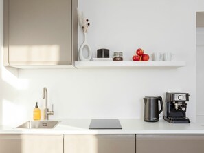 A modern kitchen with a white countertop, a coffee machine, a kettle, and a shelf with apples and jars.