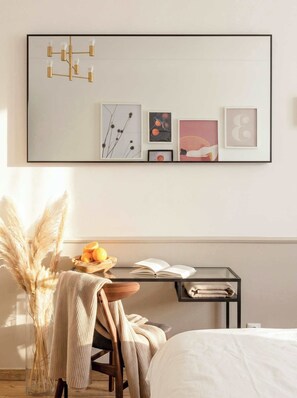 A bedroom with a bed, a bedside table with a book and a bowl of fruit, a chair with a blanket, a wall-mounted light fixture, and framed artwork.