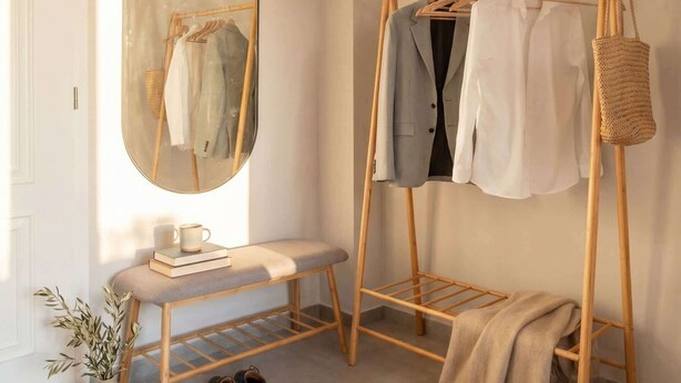 Una habitación con un perchero de madera, un espejo, un banco y una mesita con un libro y una taza.