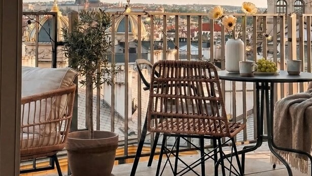 Un balcon avec des fauteuils en osier, une plante en pot et une table sur laquelle se trouvent un vase et des tasses. Une guirlande lumineuse est suspendue au-dessus.